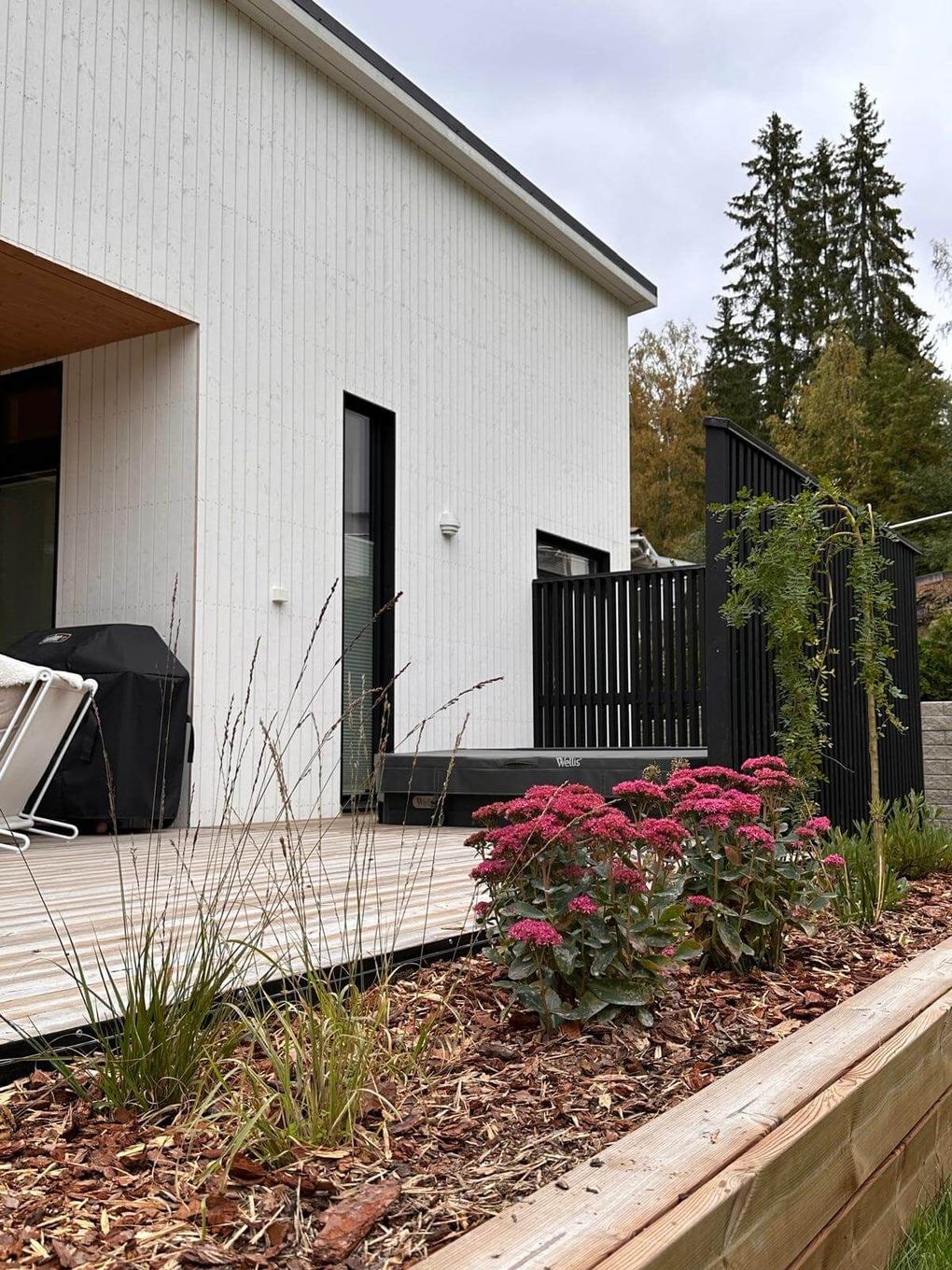 Valkoinen moderni talo, jossa on puinen terassi, poreallas, grilli ja korotettu puutarhapenkki vaaleanpunaisilla kukilla.