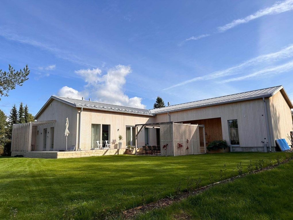 Moderni, vaalea puutalo, jossa on peltikatto ja tilava nurmikko kirkkaansinisen taivaan alla.