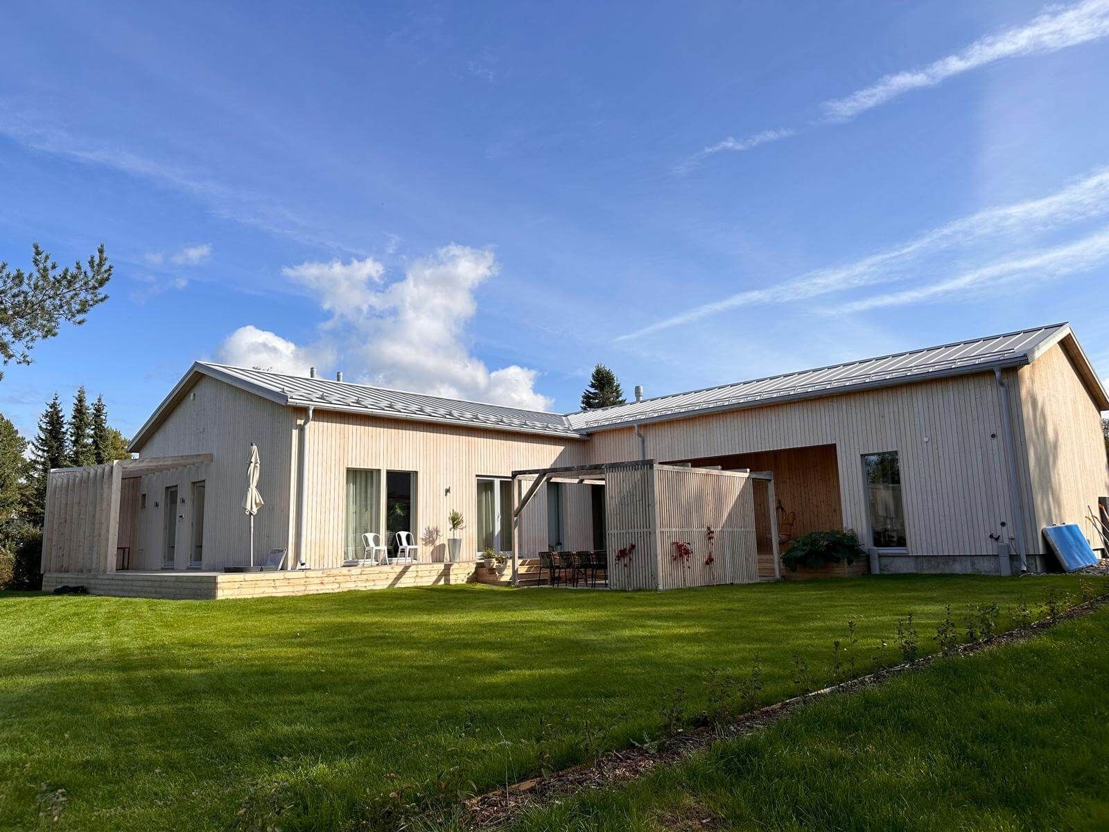 Moderni, vaalea puutalo, jossa on peltikatto ja tilava nurmikko kirkkaansinisen taivaan alla.
