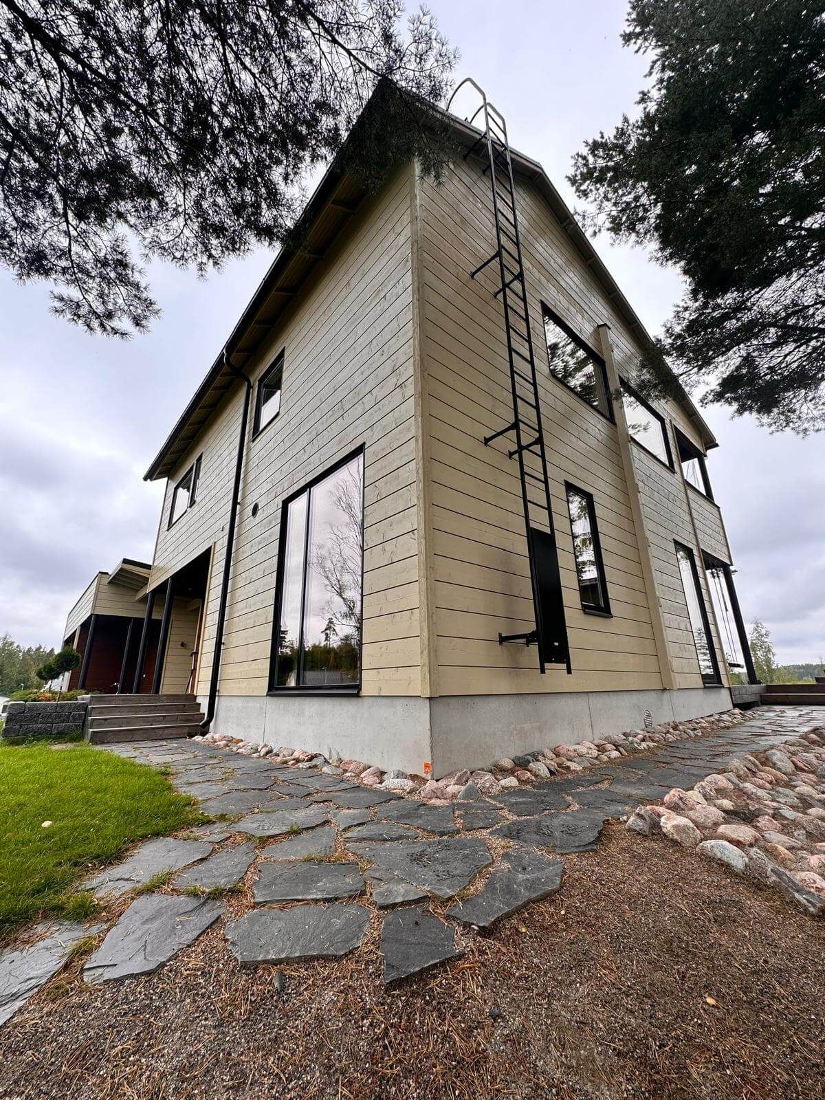 Kaksikerroksinen, vaalea puutalo matalasta kuvakulmasta, jossa on metalliset ulkotikkaat ja kivinen kävelytie.