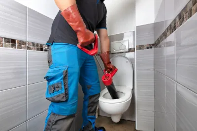 Ein Mann in blauem Overall und roten Handschuhen benutzt ein Gebläse, um eine verstopfte Toilette zu reinigen.