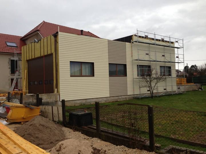 Maison à étage recouverte de bardage avec un échafaudage, un terrain en terre au premier plan et une cour clôturée