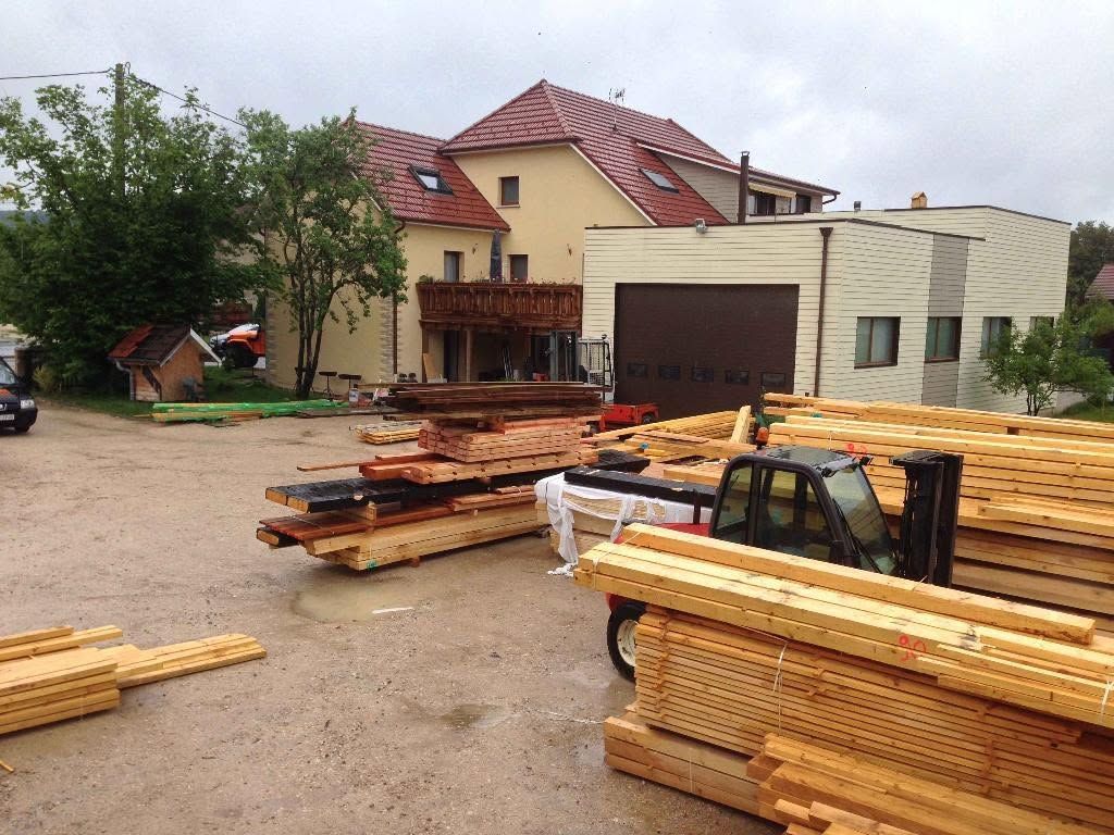 Des piles de bois et de planches à l’extérieur d’une maison en construction, avec un chariot élévateur au premier plan.