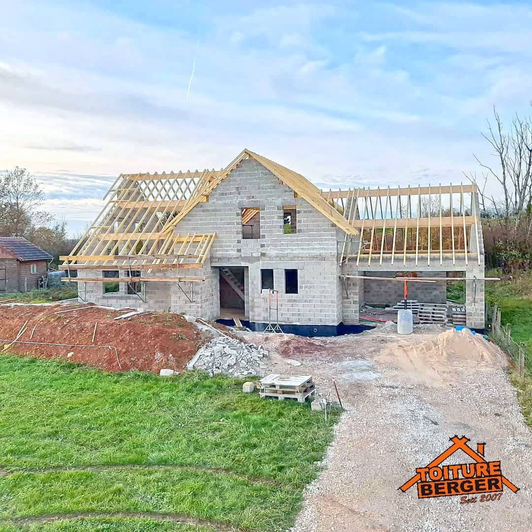 Maison en construction avec charpente de toit, murs en pierre grise et allée en gravier sur un terrain herbeux