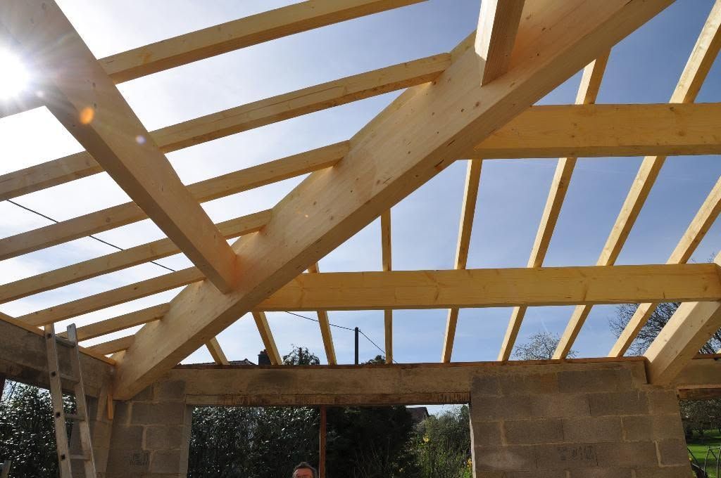 Des fermes de toit en bois en cours de construction au sommet de murs, avec le ciel visible à travers la charpente ouverte.