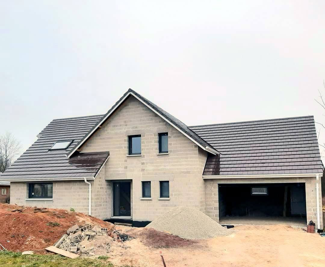 Maison de deux étages inachevée, avec un toit gris et une façade en briques, vue depuis un terrain en terre.