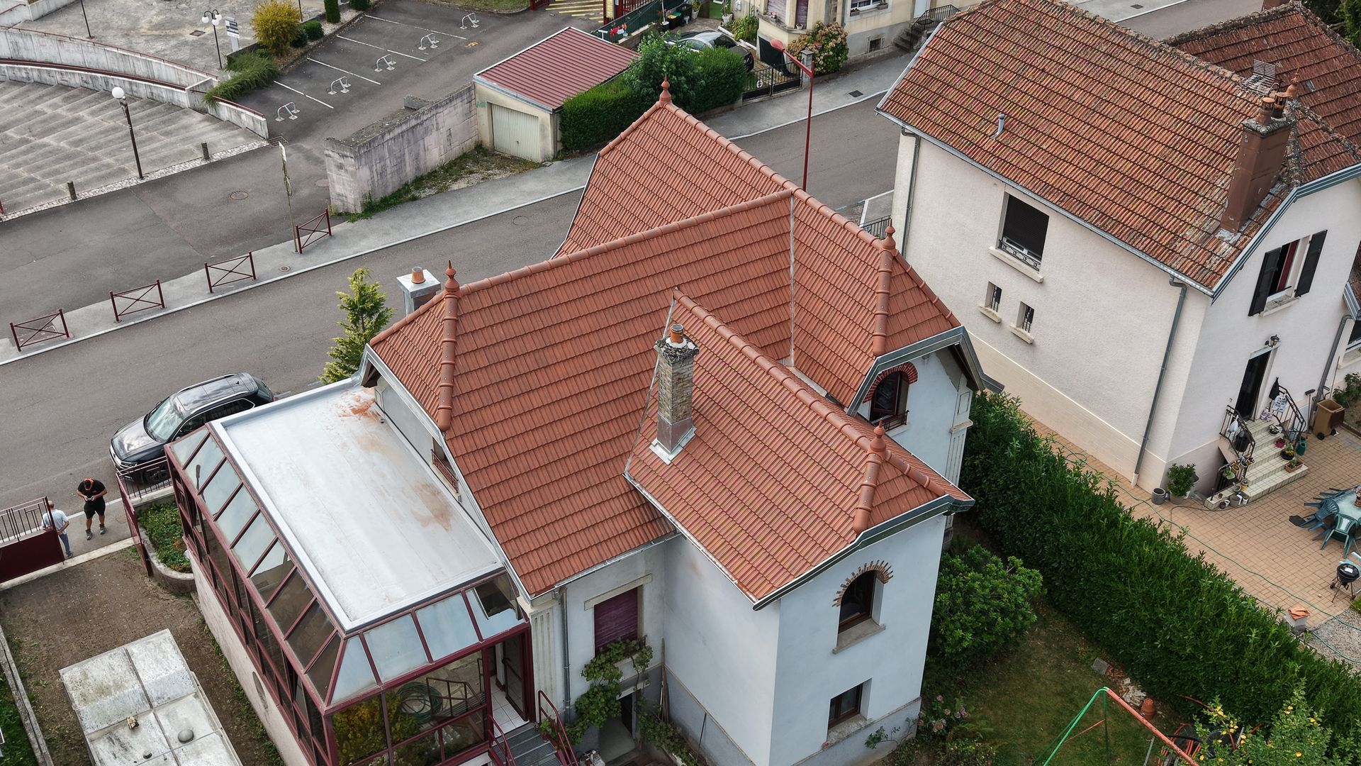Vue aérienne d’une maison blanche au toit de tuiles rouges, située au bord d’une rue et à côté de maisons voisines