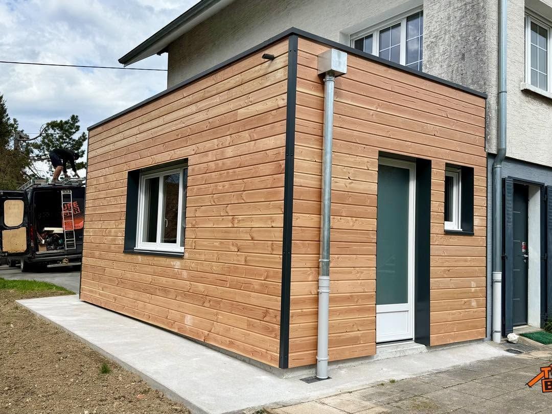 Extension de maison bardée de bois, avec des fenêtres et une porte blanche, à côté d’un bâtiment gris.