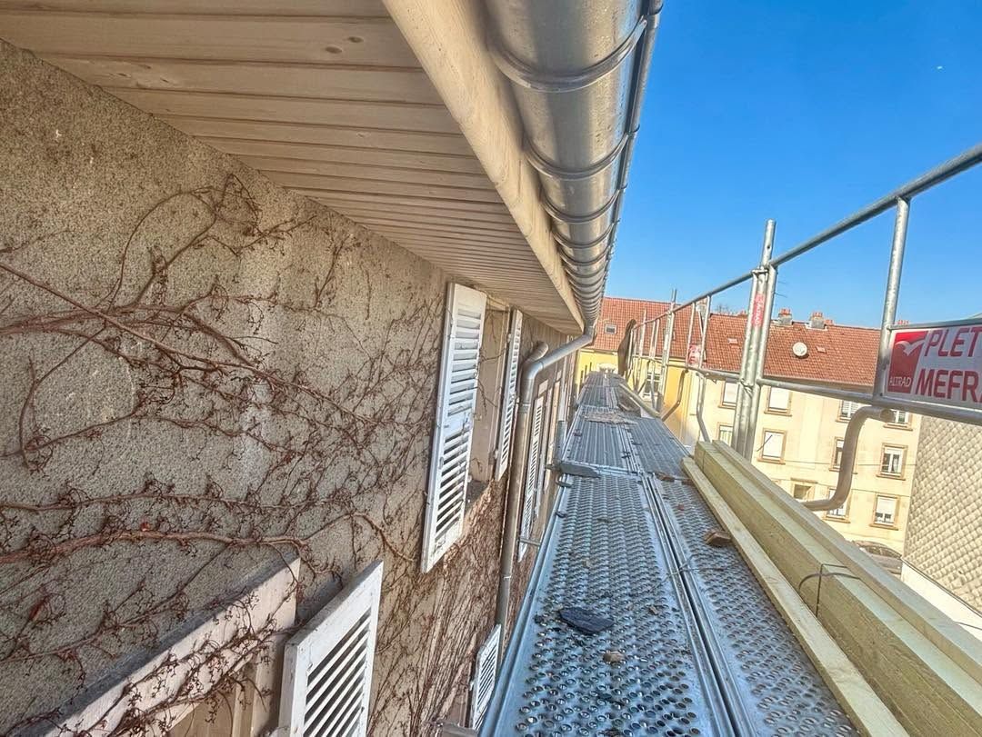 Échaffaudage le long d’un bâtiment en pierre, avec une gouttière métallique au-dessus et la rue en contrebas.