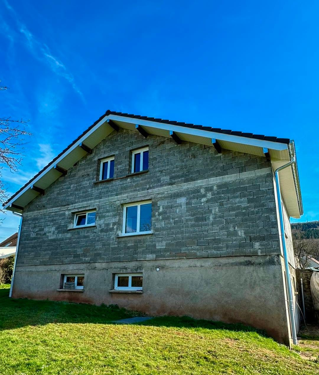 Maison en pierre grise aux petites fenêtres, avec une pelouse verte sous un ciel bleu éclatant