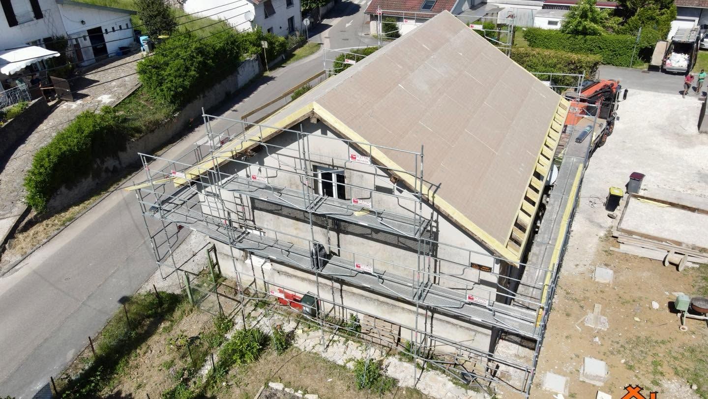 Vue aérienne d’une maison en construction, avec un échafaudage sur le toit, au bord d’une route.