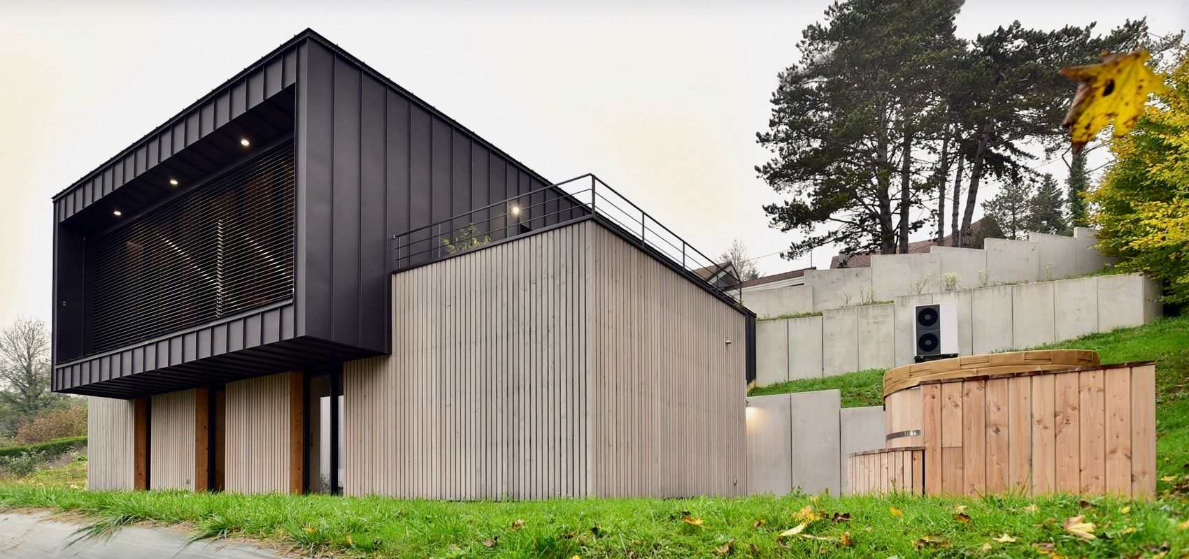 Maison moderne avec une façade supérieure sombre et un bardage inférieur en bois clair, située sur une colline verdoyante.