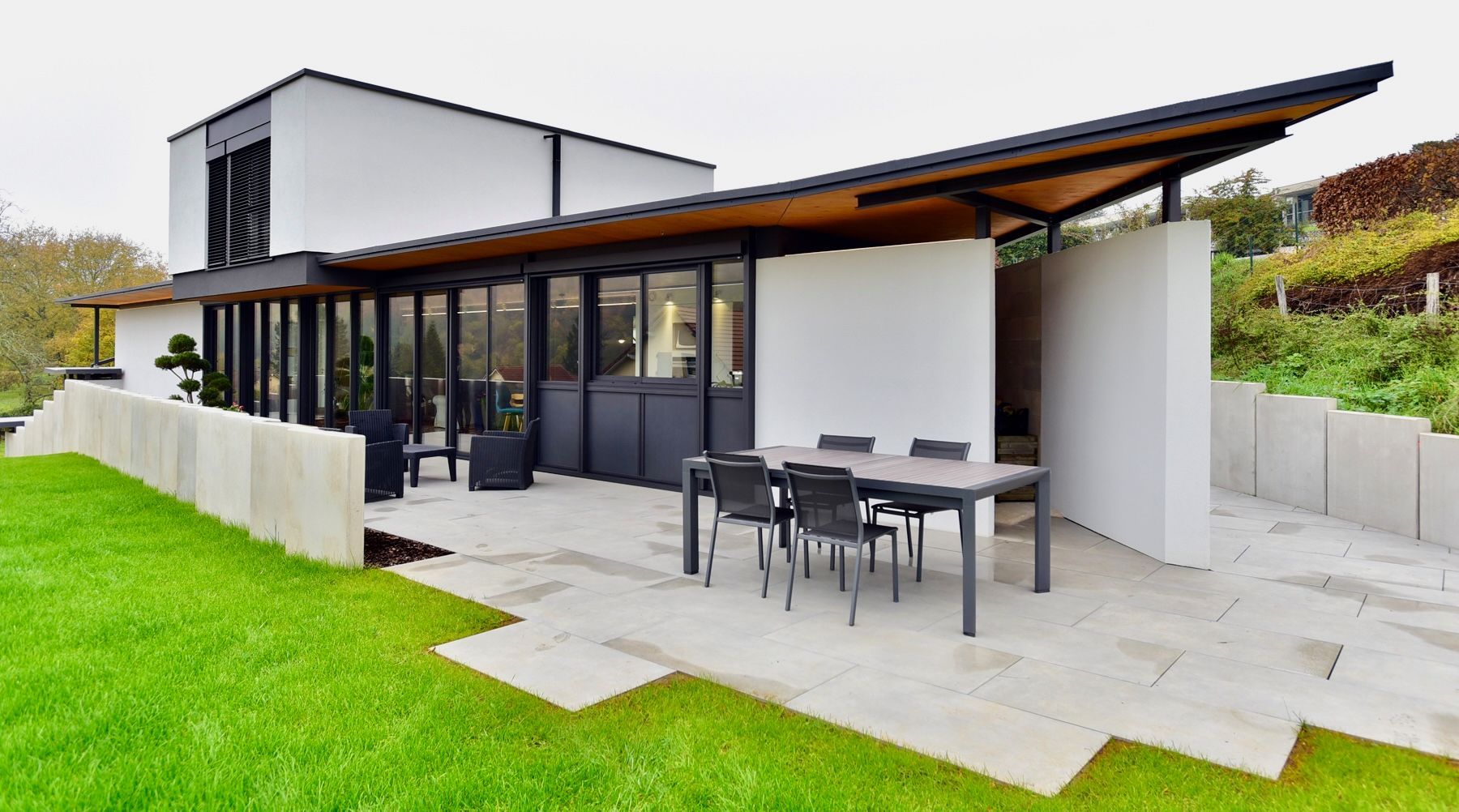 Maison moderne avec toit en pente, salon de jardin sur le patio et pelouse verdoyante à côté d’une terrasse pavée