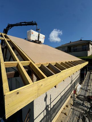 Toit en construction, avec une charpente en bois apparente et une grue qui soulève des matériaux sur fond de ciel bleu.
