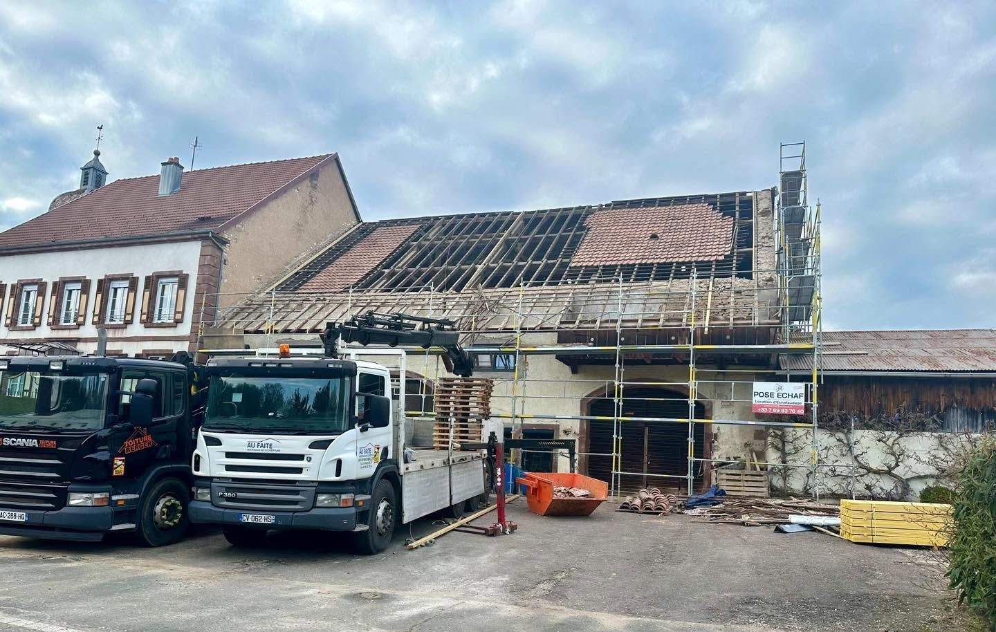 Bâtiment en rénovation avec un échafaudage, un toit endommagé et des camionnettes garées devant