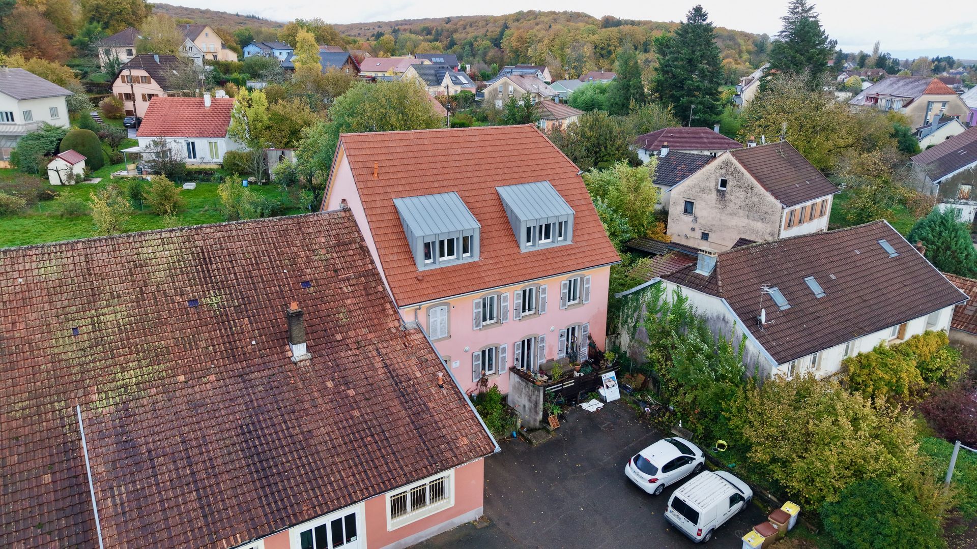 Vue aérienne de maisons aux tons pastel, aux toits rouges, et de voitures garées dans un quartier résidentiel arboré.