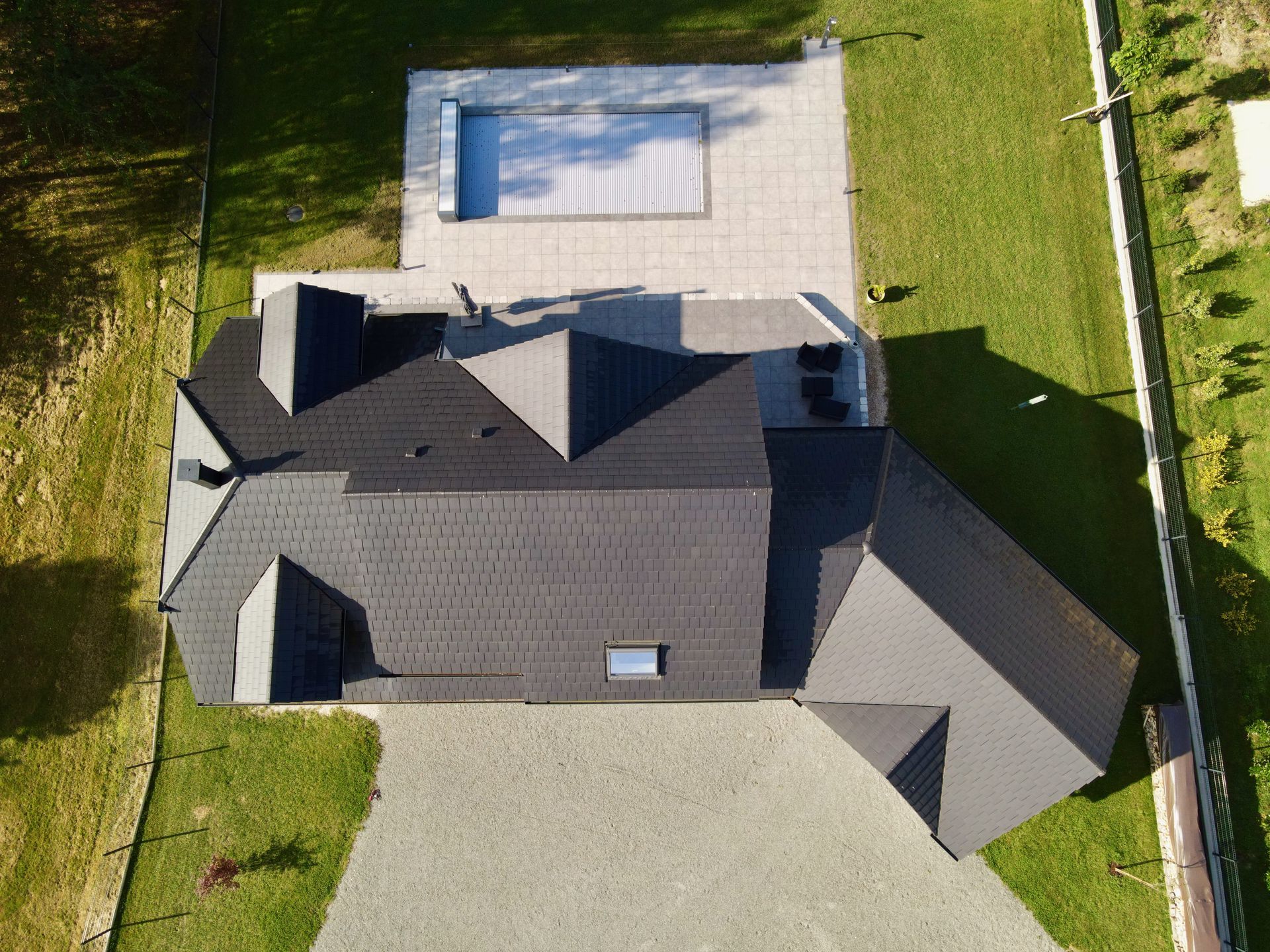 Vue aérienne d’une maison au toit sombre, avec une allée claire et une piscine dans le jardin.