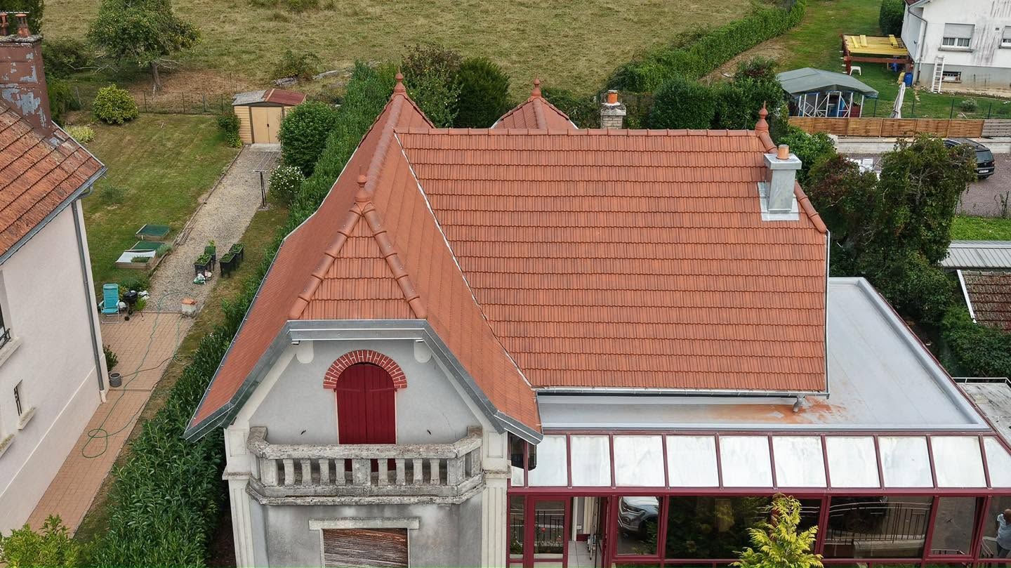 Vue aérienne d’une maison au toit rouge avec une lucarne, une terrasse à l’avant et de la verdure tout autour.