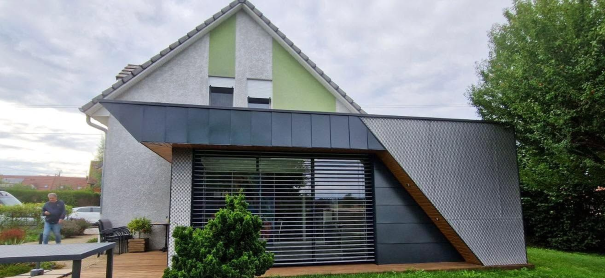 Maison moderne au toit en pente, avec une grande fenêtre à l’avant et une façade gris-vert sous un ciel nuageux