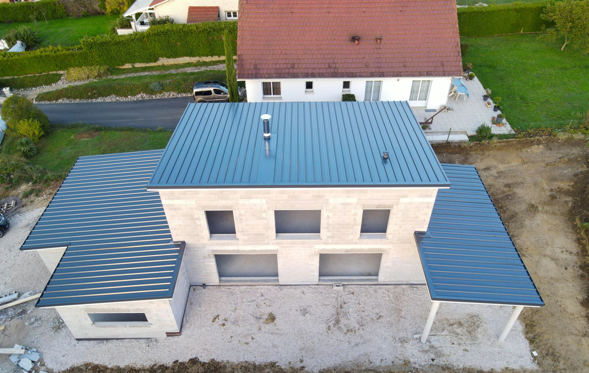 Vue aérienne d’une maison à étage, avec un toit bleu et un garage attenant, près d’un bâtiment blanc