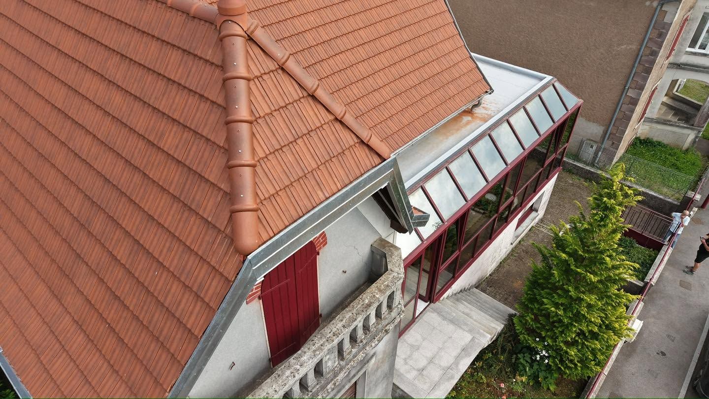 Vue aérienne d’une maison au toit rouge, avec une véranda vitrée et un jardin étroit en bordure de rue.
