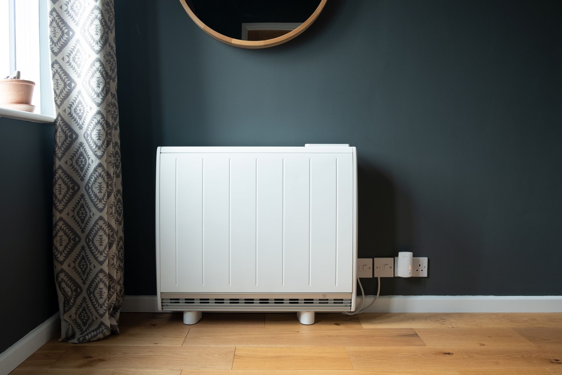 Radiateur sous un miroir rond dans une pièce aux murs sombres, avec des rideaux à motifs et un parquet.