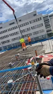 Rakennustyömaa, jossa työntekijöitä neonvaloihin pukeutuneina turvavarusteisiin raudoitusalustalla kallistuneen rakennuksen vieressä