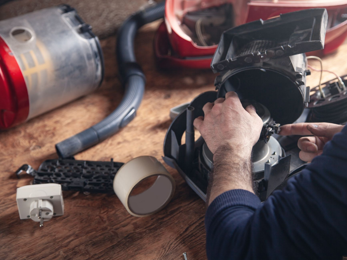 Une personne répare un aspirateur noir avec des outils, entourée de pièces détachées.