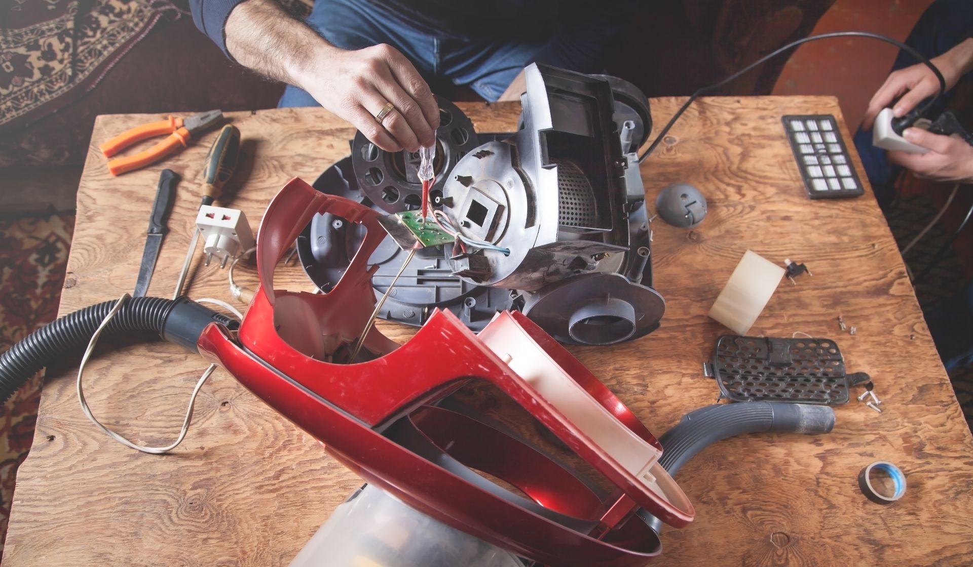 Des mains réparent un aspirateur rouge sur un établi en bois, des outils éparpillés autour.