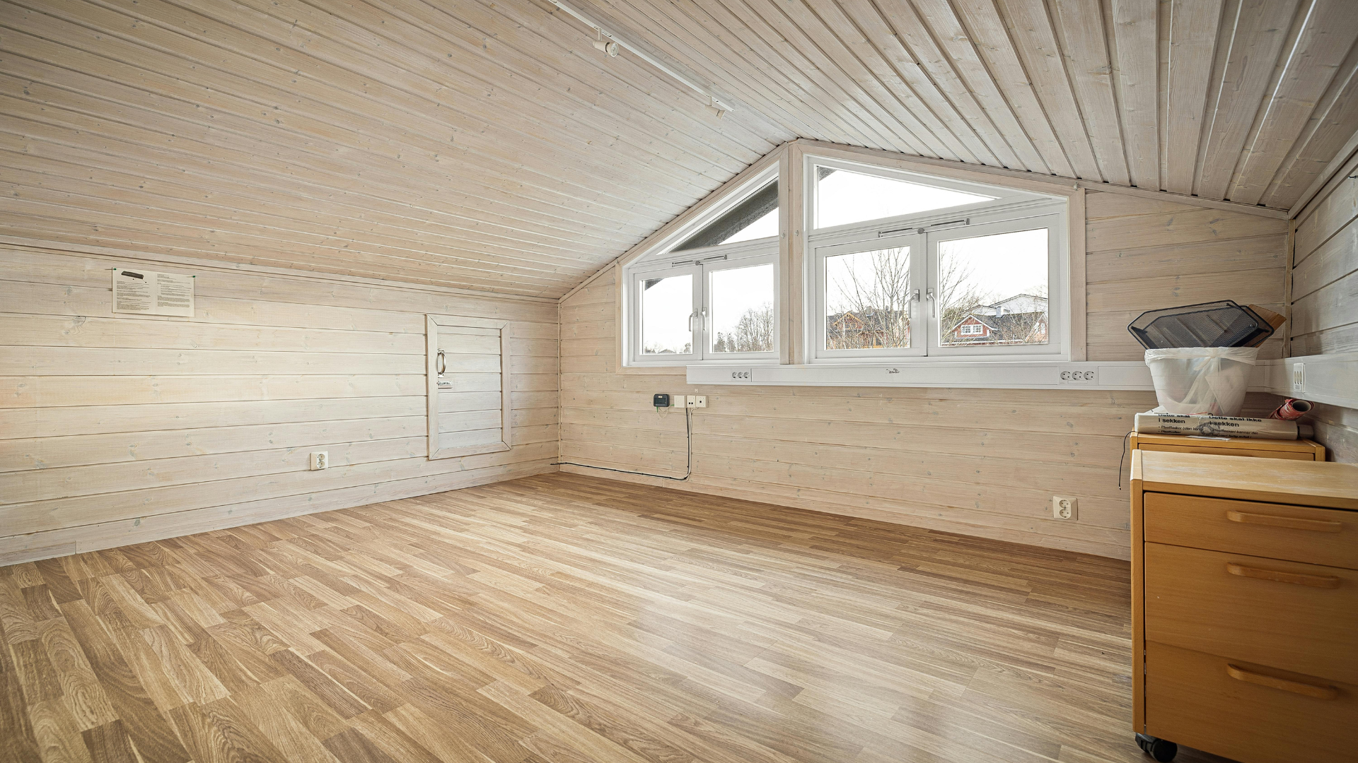 Chambre mansardée vide avec parquet, murs et plafond lambrissés blancs, grande fenêtre.