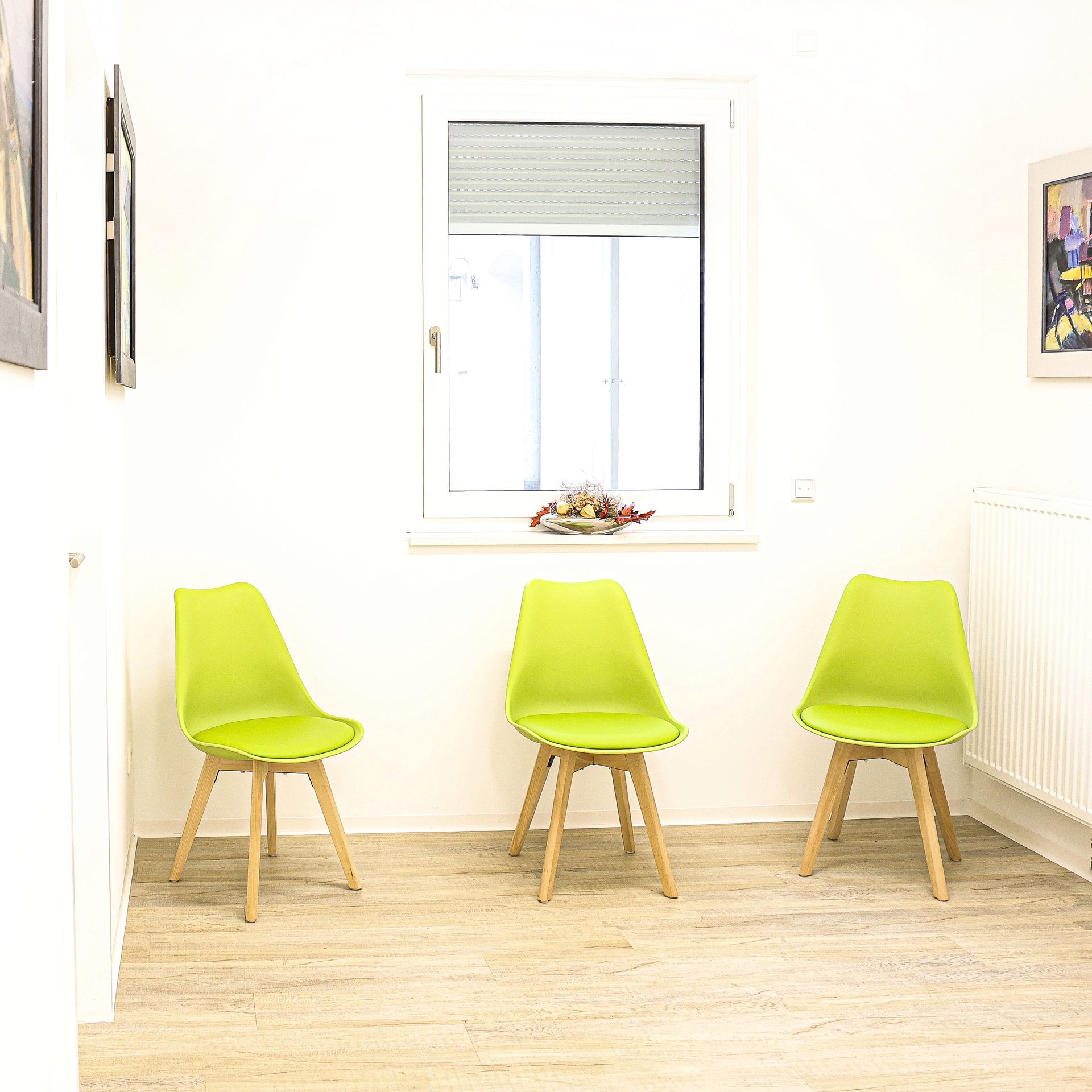 Drei grüne Stühle in einem hell erleuchteten Raum, in der Nähe eines Fensters mit Jalousien, vor einer weißen Wand.
