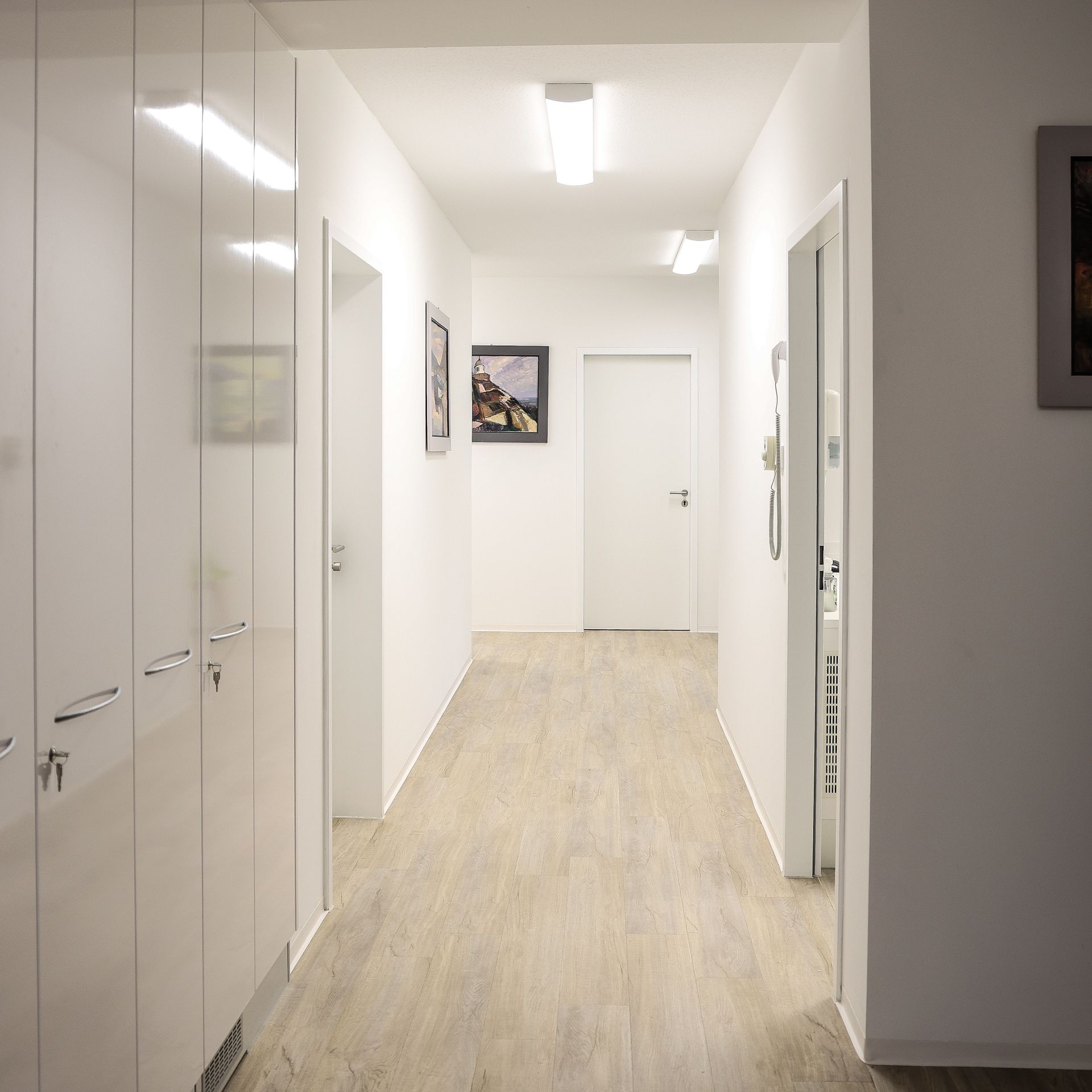 Ein strahlend weißer Flur mit hellem Bodenbelag, geschlossenen Türen und Kunstwerken an den Wänden.