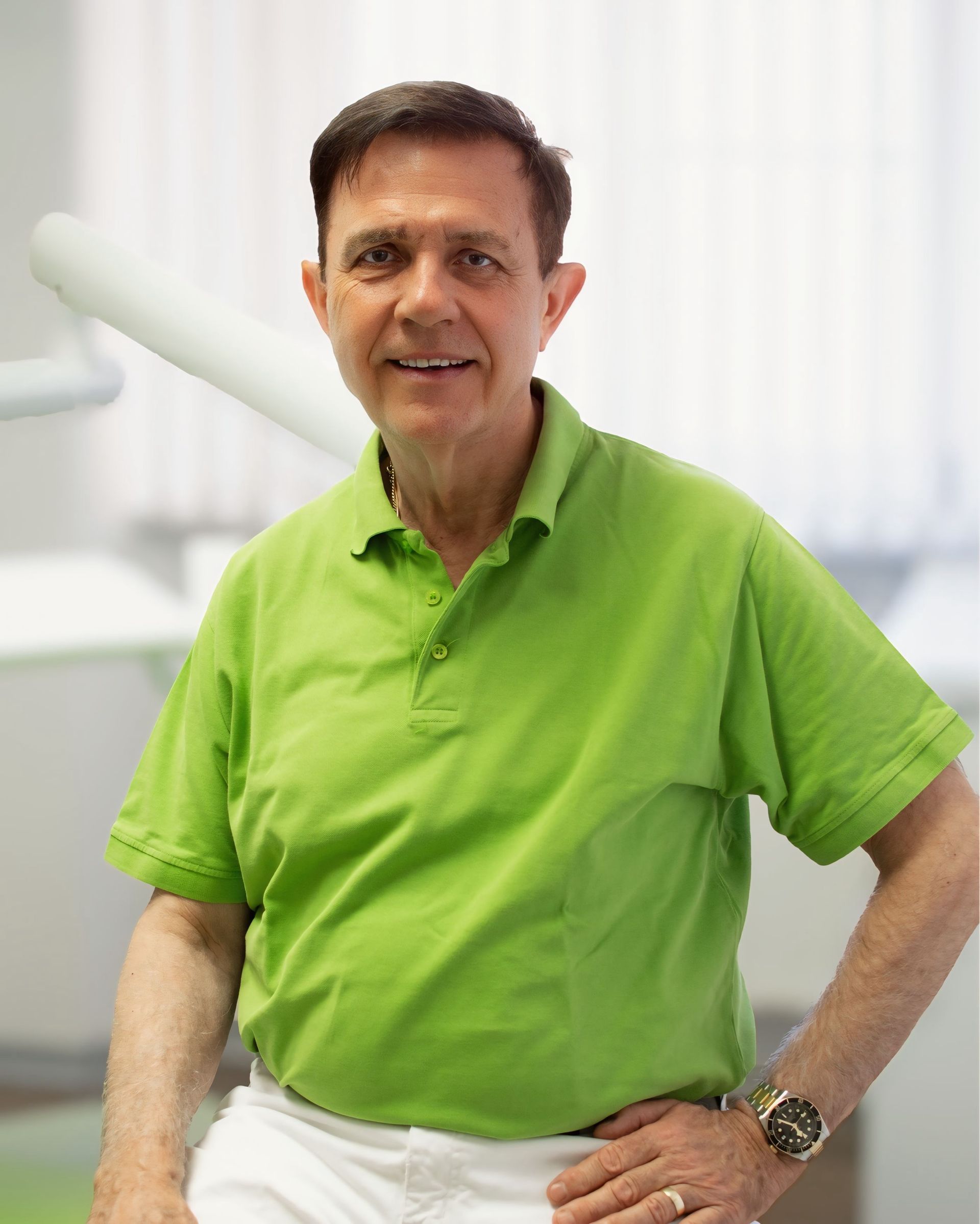 Mann im grünen Poloshirt, lächelnd, Hand in die Hüfte gestemmt, im Hintergrund eine Zahnarztpraxis.