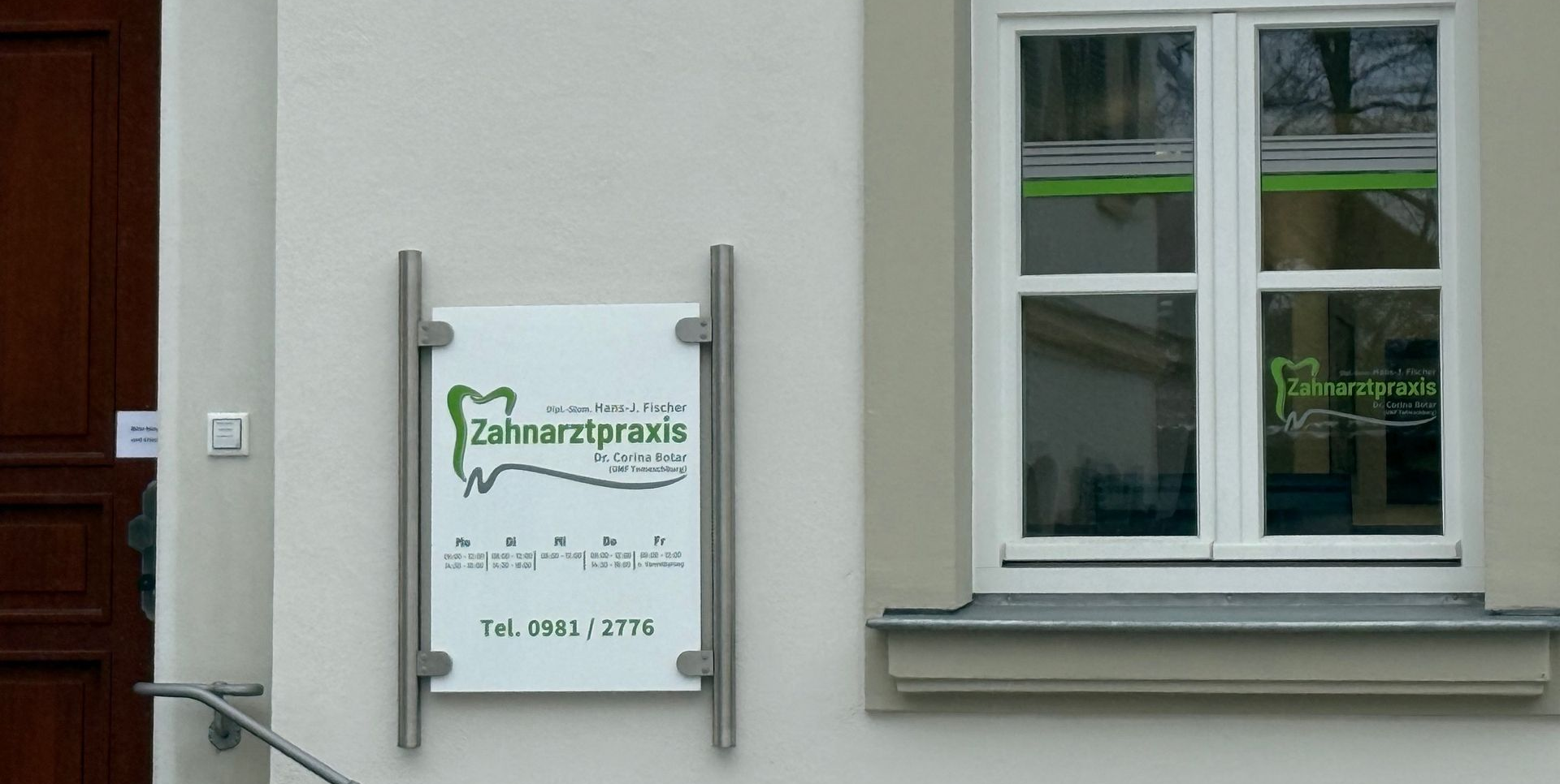 Zwei weißgerahmte Fenster an einem beigen Gebäude mit einem Schild auf der linken Seite.