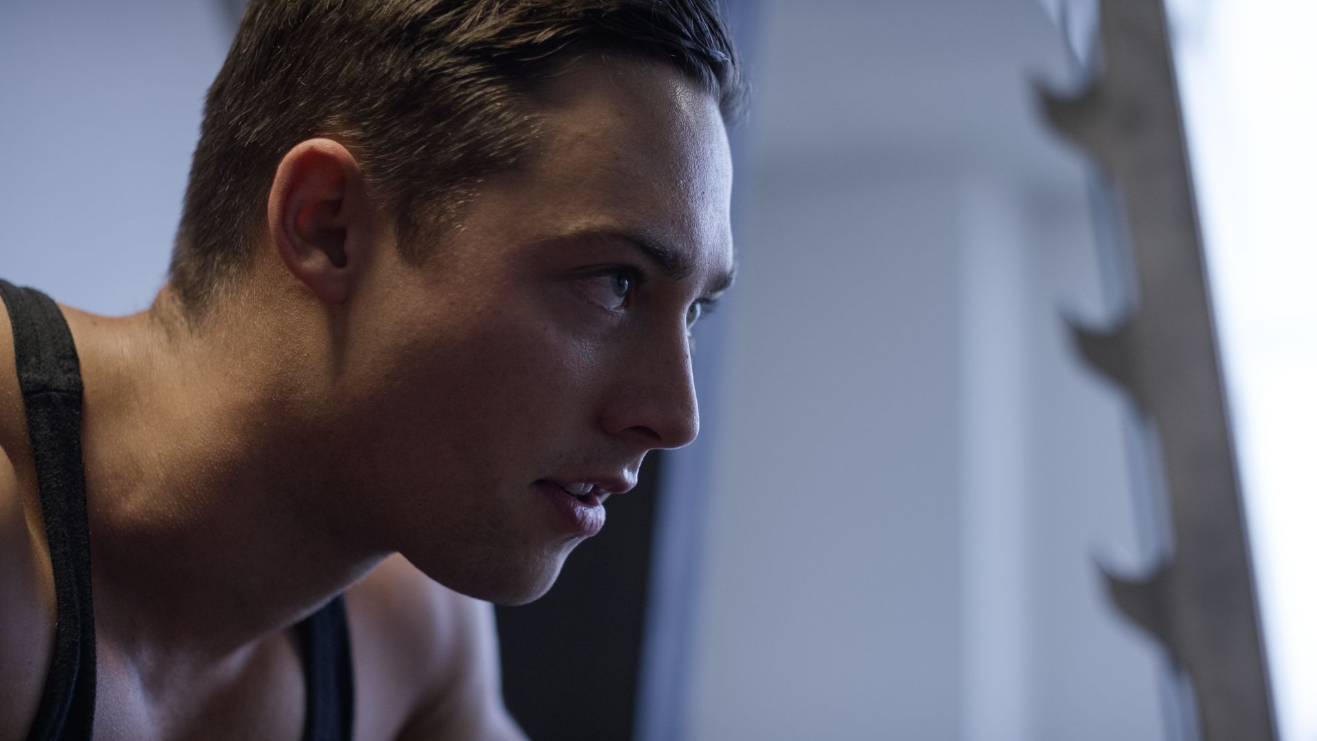 Gros plan sur une personne concentrée, vêtue d'un débardeur sombre, penchée en avant dans une salle de sport faiblement éclairée.