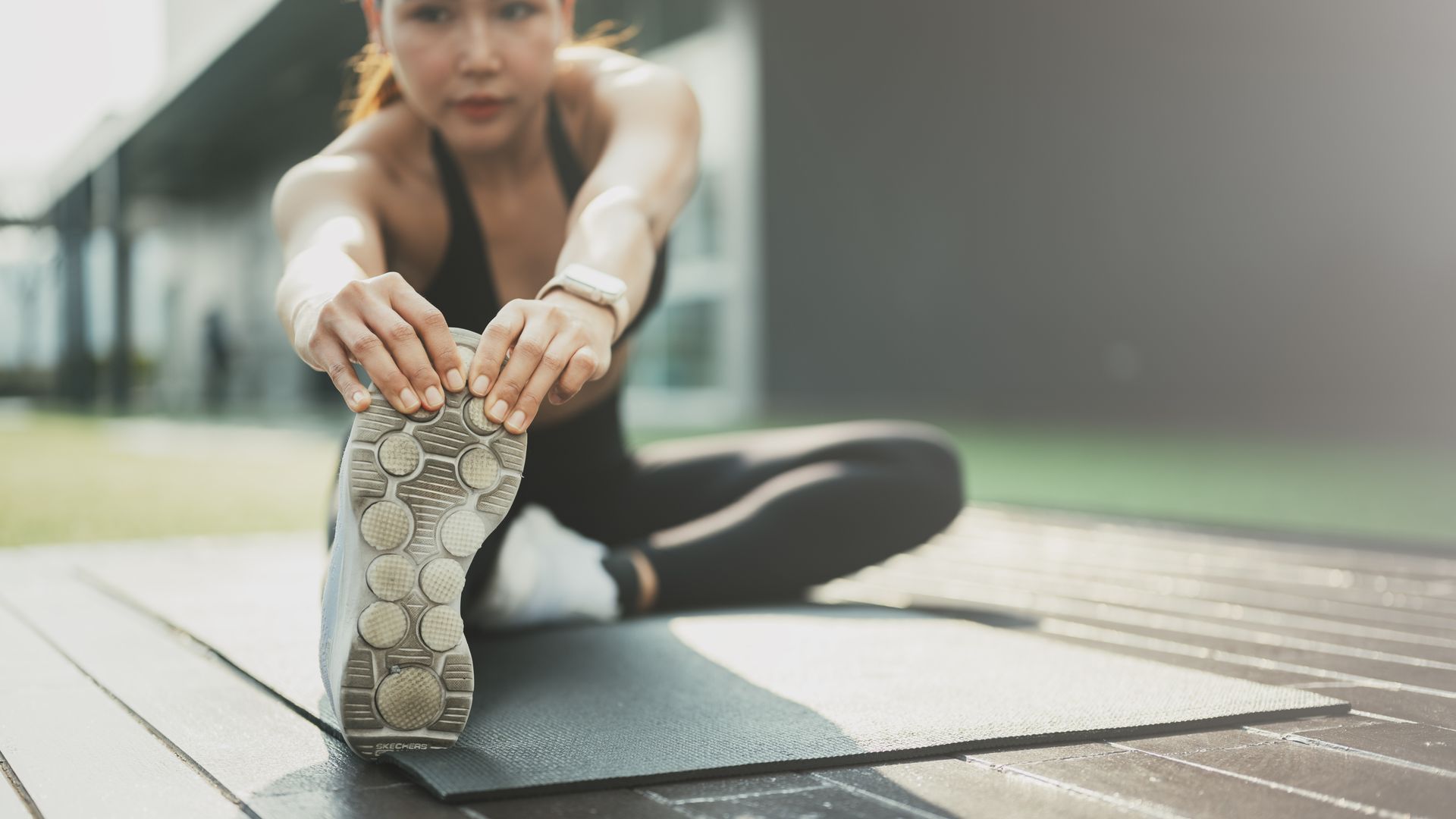 Une personne étirant une jambe sur un tapis à l'extérieur, tendant la main vers la semelle d'une basket dans un parc flou.
