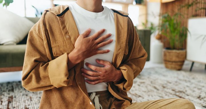Une personne assise en tailleur sur un tapis, se tenant la poitrine et l'abdomen, dans un salon confortable.