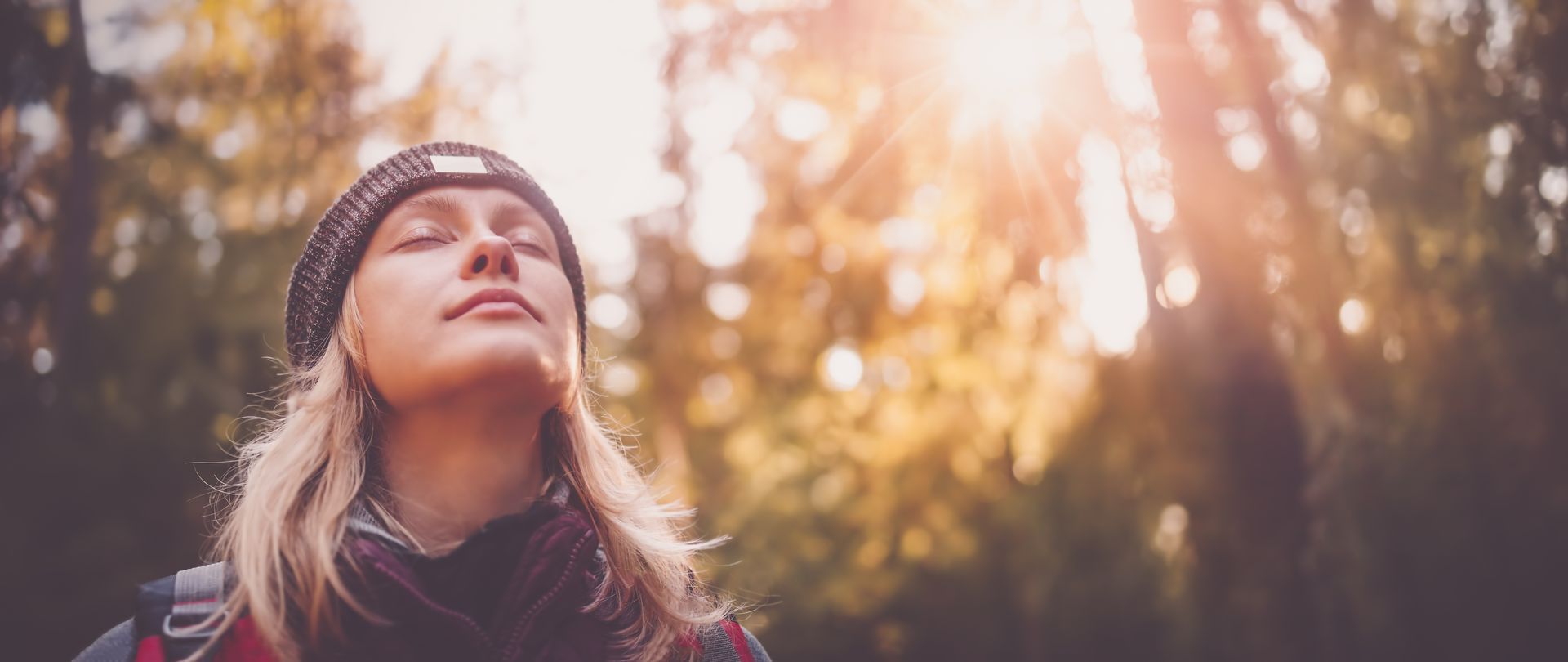 Une femme coiffée d'un bonnet en laine lève les yeux vers une forêt ensoleillée, où une douce lumière dorée filtre à travers les arbres.