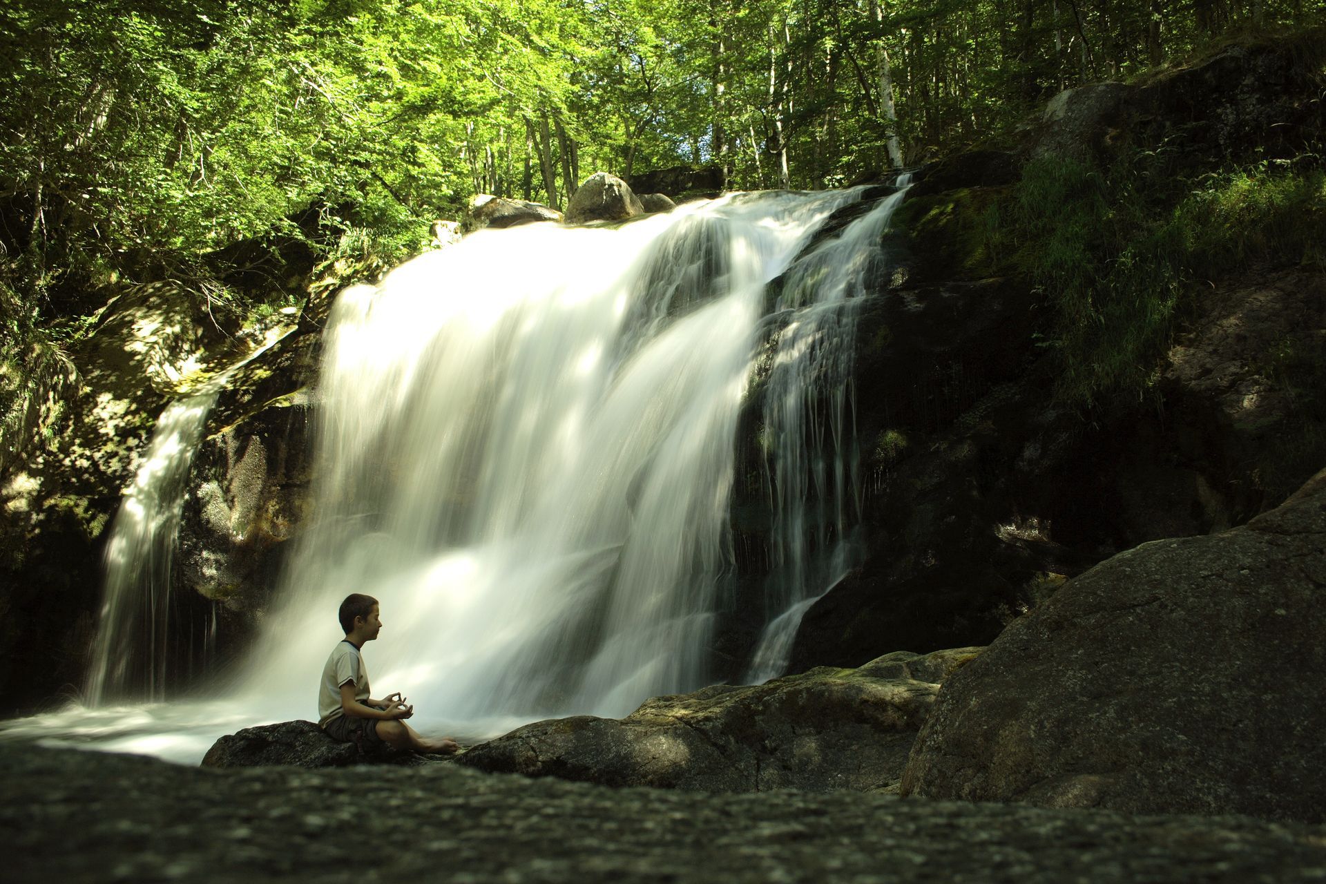 Personne assise près d'une cascade en forêt, baignée d'une lumière du soleil tamisée.