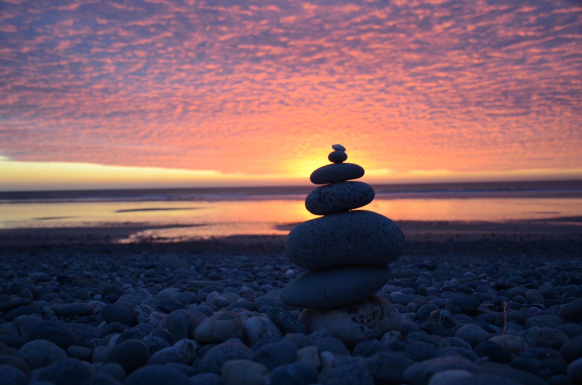 Des pierres empilées se découpent sur une plage rocheuse au coucher du soleil, sous un ciel orangé et rose.
