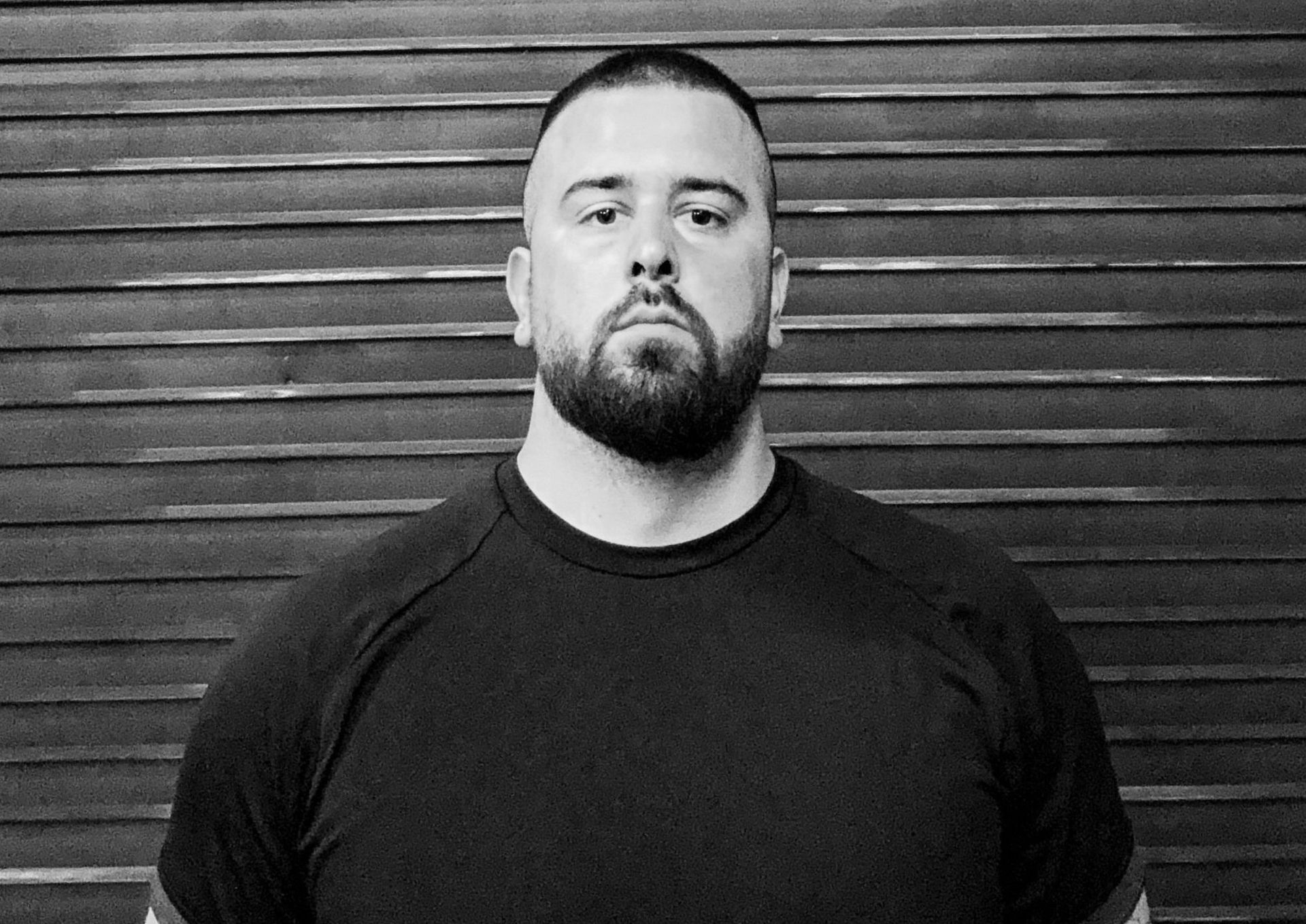 Man with a beard and shaved head in a dark shirt, standing in front of a shuttered door.
