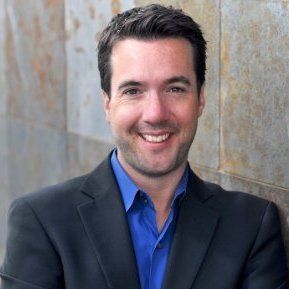 Man in blue shirt and blazer smiles, leans against stone wall.