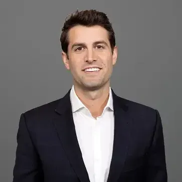 Man in a suit smiles, white shirt, against a gray background.