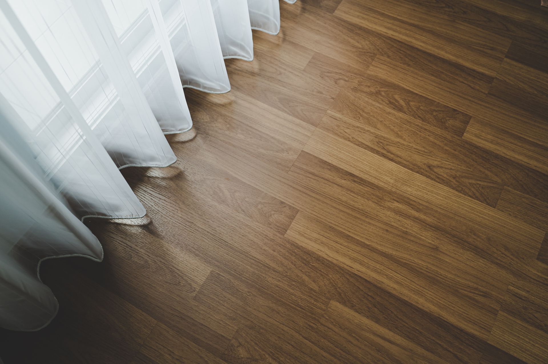 Des voilages blancs à côté d’un parquet brun baigné de soleil, avec de douces ombres.