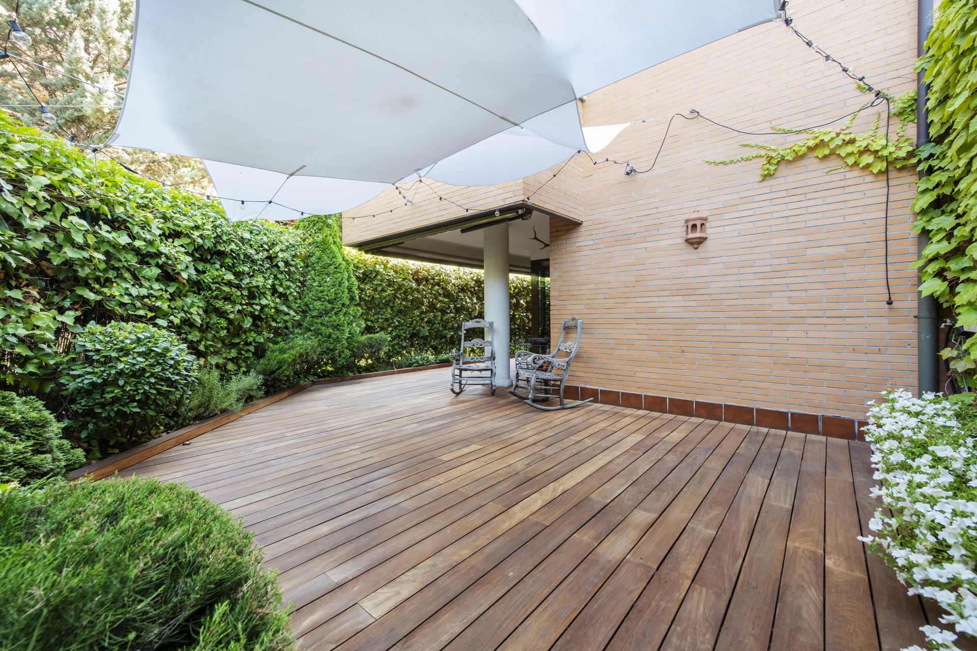 Terrasse en bois dans un jardin, avec des chaises de patio, des voiles d’ombrage blanches et une végétation luxuriante.