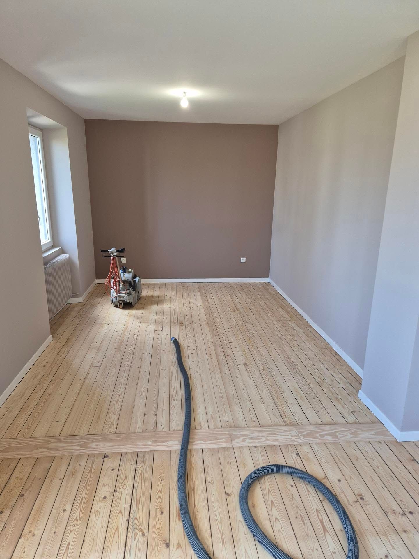 Pièce vide avec un parquet brut, des murs beiges et une ponceuse pour parquet sur le sol.