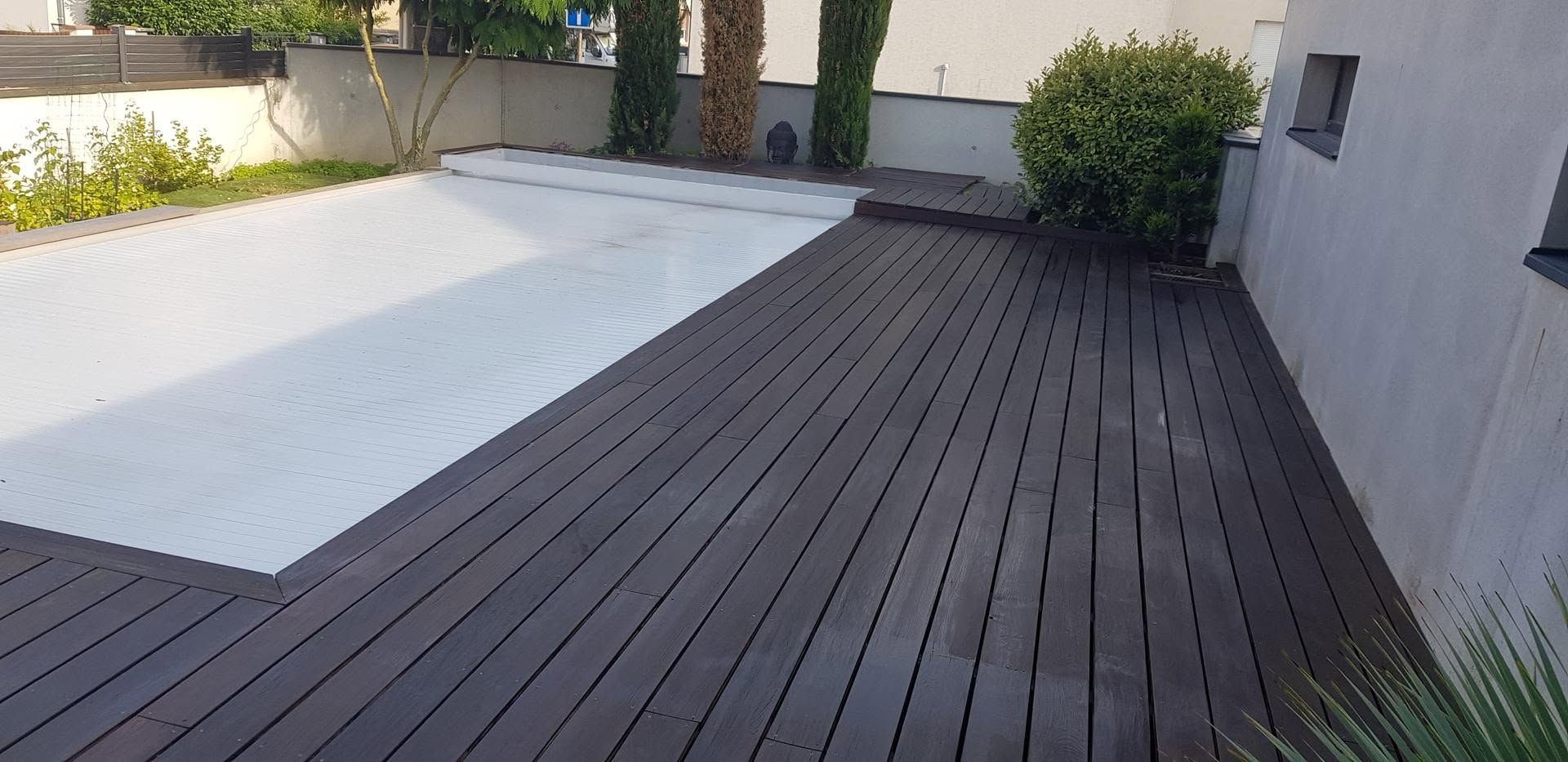 Piscine recouverte d'une bâche blanche dans un jardin, avec une terrasse en bois foncé et une végétation environnante.