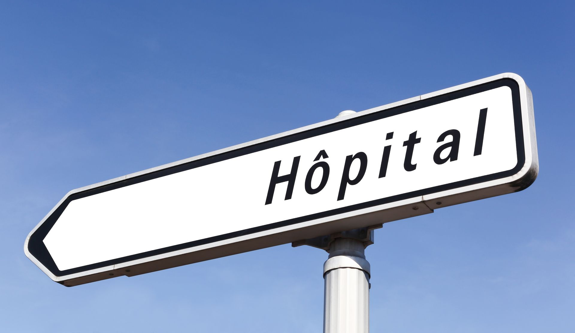 Panneau routier pointant vers la gauche, avec le mot Hôpital sur un panneau rectangulaire blanc, ciel bleu en arrière-plan.