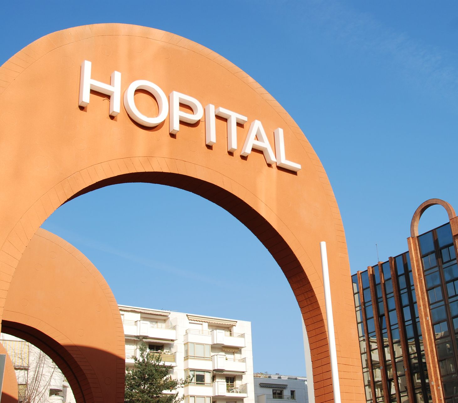 Arche orange avec l'inscription HÔPITAL, ciel bleu et bâtiments.