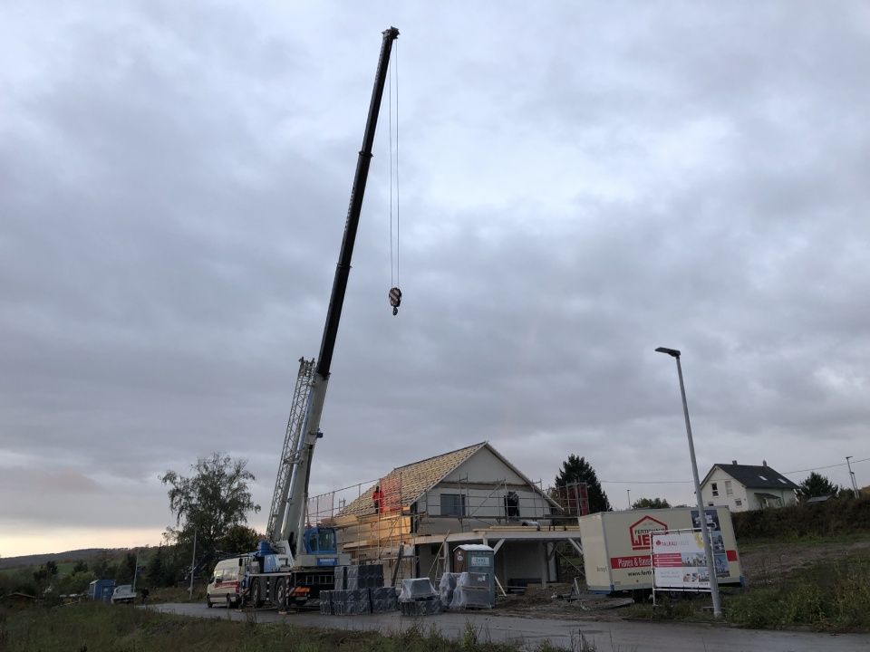 Ein großer Kran hebt ein Stück Holz vor einem im Bau befindlichen Haus an.